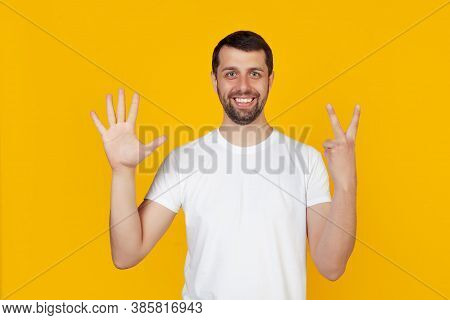 Modern Young Man With A Beard In A White Tank Top Shows Number Seven With Fingers On Hand, Smiling C