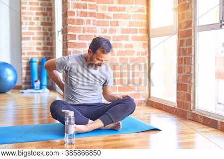 Middle age handsome sportman sitting on mat doing stretching yoga exercise at gym Suffering of backache, touching back with hand, muscular pain