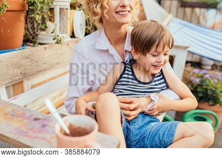 Blond Curly Woman Hugging, Caressing And Playing With Her Child Outdoor At Open Ait Terrace Or Backy