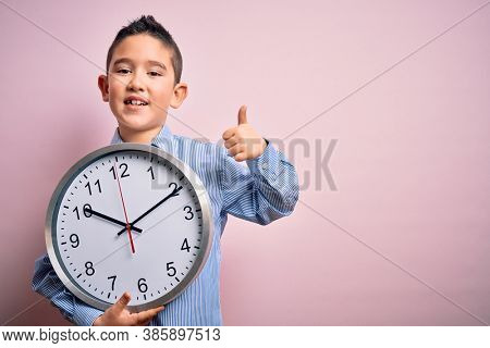 Young little boy kid holding big minute clock over isolated pink background happy with big smile doing ok sign, thumb up with fingers, excellent sign