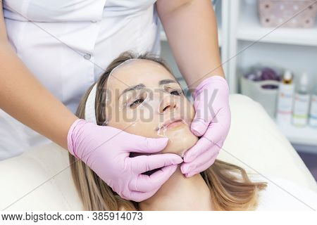 Cosmetologist Applies A Hydrogel Mask To A Young Woman On Her Face. The Procedure By A Beautician. R