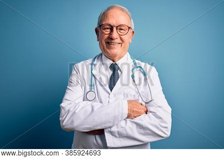 Senior grey haired doctor man wearing stethoscope and medical coat over blue background happy face smiling with crossed arms looking at the camera. Positive person.