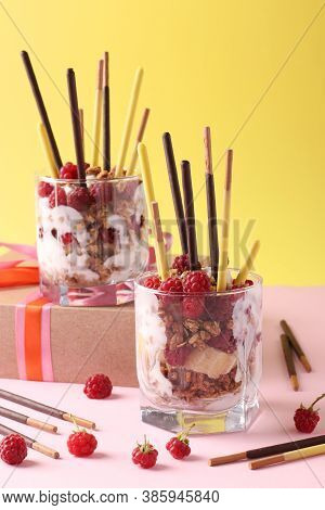 Two Glasses With Chocolate Granola, Banana And Raspberry, Decorated With Sticks Pocky On Yellow And 