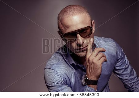 Suave handsome man in sunglasses with his hand to his cheek looking at the camera with a serious expression, on grey with copyspace