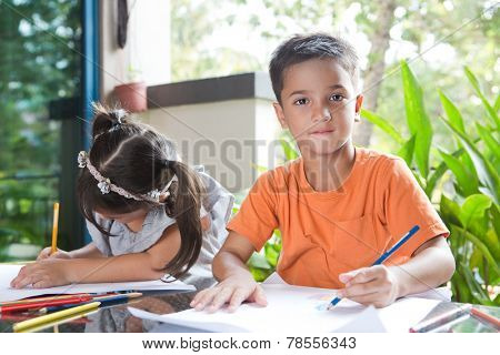 Cute little pan asian boy smiling while holding a color pencil sitting next to his younger sister engross in her coloring activity in home environment
