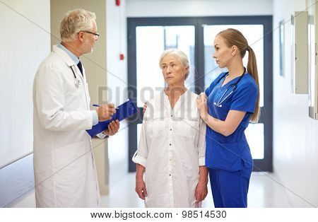 medicine, age, health care and people concept - male doktor with clipboard, young nurse and senior woman patient talking at hospital corridor