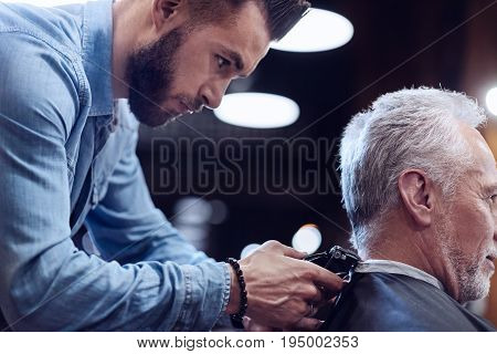 Professional barbershop. Nice handsome male hairdresser using a hair cutting machine and focusing on his work while standing behind his client