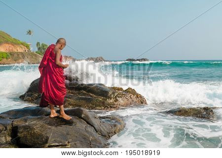 A young Buddhist monk stands on a dark stone in the ocean