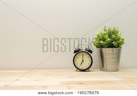 black vintage alarm clock times at 7 o'clock morning and green plant tree in silver pot small minimal decorative on wooden desk and white wall background vintage tone retro style