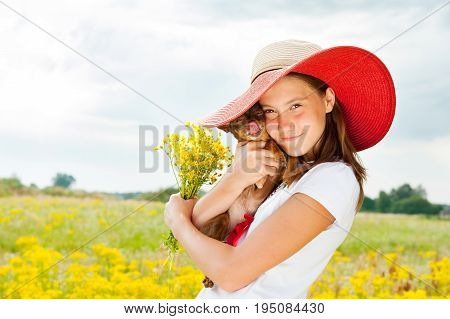 Young cheerful teenage girl hugging her little dog in field with bouquet of yellow flowers. Multicolored summertime outdoors horizontal image.