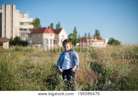 cool little boy with a grimace on his face stands in a field on the background of houses
