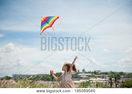 happy small girl with long hair holding flying colorful kite in the field on summer sunny day on sky and town backgound