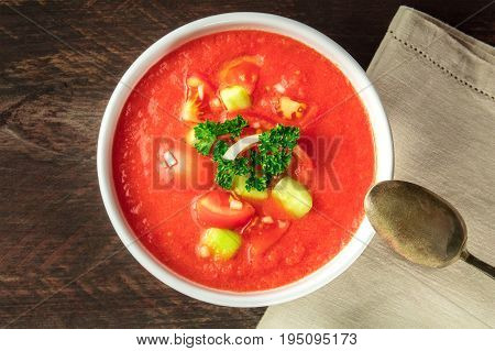 A bowl of gazpacho soup with slices of tomato, cucumber, and onion, with parsley leaves, shot from above on a dark rustic texture with a spoon and a place for text