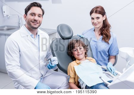 Little Boy Sitting In Chair With Smiling Dentists In Dental Clinic