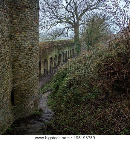 The old fortress wall and a fragment of the tower, green with time. City of Bergues, France. In the foreground is a overgrown hillside and a stone staircase.