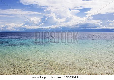 Seaside landscape with turquoise water and tropical island. Sea view with distant island. White fluffy clouds above blue ocean. Sea and sky minimal photo background. Idyllic seashore with white sand