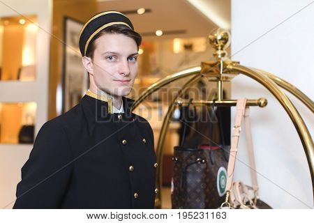 Smiling man in bellboy uniform standing with luggage cart and looking at camera