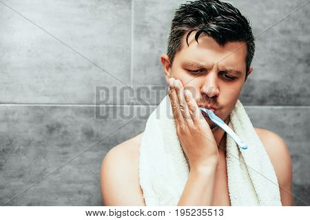 Toothache. Frustrated young handsome man touching his cheek while brushing teeth in bathroom. sensitive teeth and gums. Dental hygiene, health care, beauty and people concept.