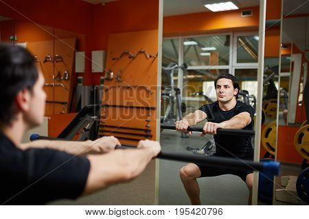 Young sporty man in the black sportwear with barbell doing squats in gym n front of the mirror. Athlete man in the t-shirt and shorts. Healthy lifestyle. Power and energy.