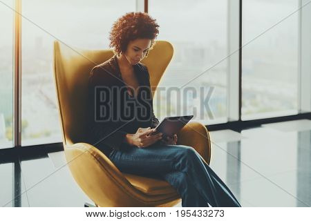 Young cute Brazilian probationer using digital tablet to prepare for interview while sitting in yellow armchair pensive afro american girl working on digital pad in office settings near huge window