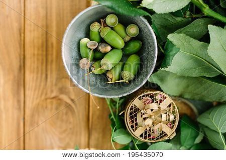 environment, nature, countrylife concept - flat-lay of little bowl with fresh green acorns, leaves of rose bushes and small cardboard gift box with pink dried flowers on wooden table