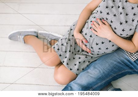 pregnancy, family love and happiness concept - closeup on round abdomen of young healthy lady, future mother and father sit side by side on white wooden floor, expecting child birth happiness.