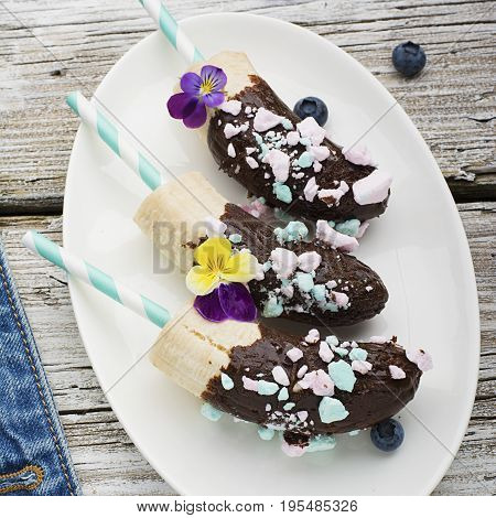 Chocobanana Light summer refreshing dessert made from frozen banana Eskimo in a glaze made of dark chocolate and colored meringue on a cocktail tube on a light background. Top View.