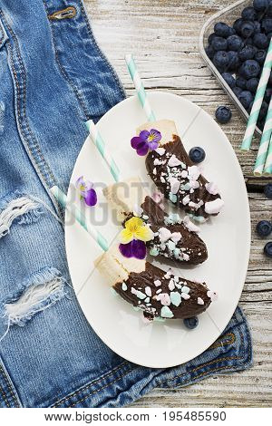 Chocobanana Light summer refreshing dessert made from frozen banana Eskimo in a glaze made of dark chocolate and colored meringue on a cocktail tube on a light background. Top View.