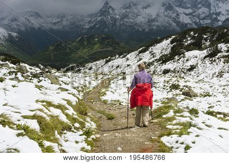 A female senior hiker wearing a red coat hiking through snow in summer on the way to the Zeinisjoch in the Paznaun Valley near Galtur in Austria.