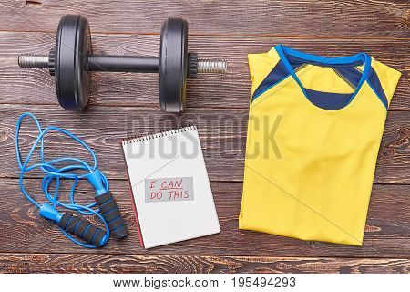 Athletic clothes and sport equipment. Message, jumping rope, dumbbell, t-shirt on wooden background. Be stronger and healthier with gym workout.