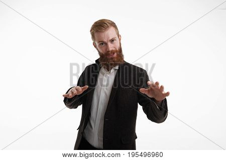 Bearded businessman with attitude wearing formal suit standing at white wall gesturing with hands as if saying Calm dowm or Relax. Negative human emotions facial expressions signs symbols