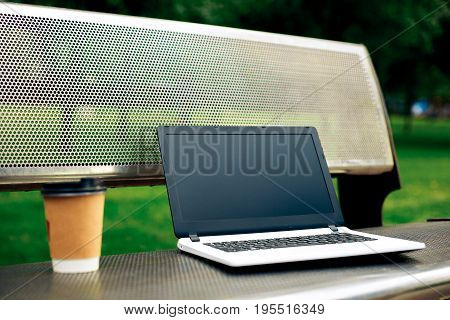 Mockup image of laptop with blank black screen and coffee cup on metal bench in nature outdoor park. Freelance work.