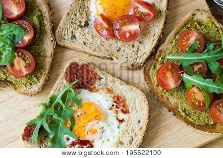 Toasts with healthy bread with sesame seeds egg cherry tomato arugula spinach pesto and tomato sauce on wooden board. Breakfast meal