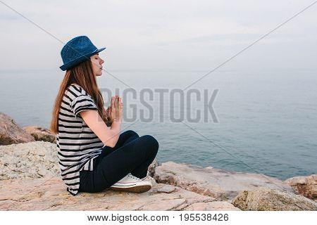 Young beautiful girl in a hat practicing yoga and meditating on stones next to the sea. Recreation. Relaxation.