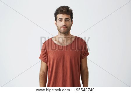 Portrait of stylish bearded guy with trendy haircut wearing casual red T-shirt looking with his brown eyes at the camera. Young handsome man having pleased look. People and emotions concept.