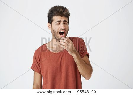 Yawning young fellow with beard and trendy hairstyle tired of work and holding his hand behind his mouth. Student wearing red T-shirt got bored of lectures.