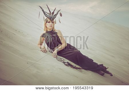 Woman with horns on her head lying on the sand in the desert.