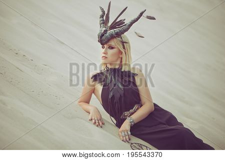 Beautiful woman with animal horns on her head lying on the sand in the desert.