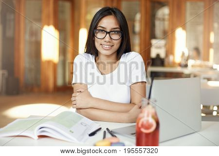 Portrait of assian woman smiling working on laptop in cafe or bright coworking space wearing glasses.