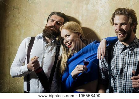 Girl with liqueur glass smiling with macho and hipster holding bottle of wine. Men and woman with alcoholic drinks on beige wall. Alcohol convives and alcoholism. Unhealthy lifestyle and bad habits