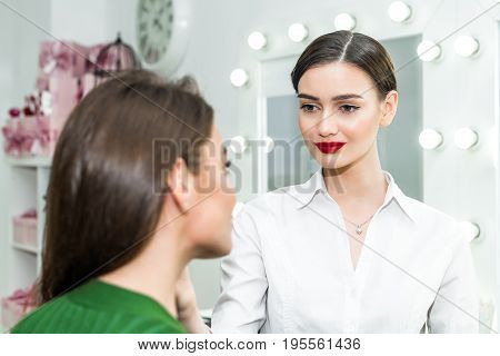 Attractive brunette girl primp at the mirror with lamps in the beauty studio. A happy satisfied customer of make-up and hairstyle service.