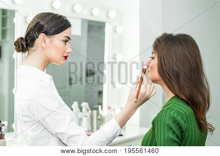 Attractive brunette girl primp at the mirror with lamps in the beauty studio. A happy satisfied customer of make-up and hairstyle service.