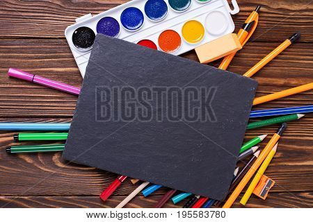 Different stationery - pencils pen note erazer paint and empty slate board on wooden background. Top view. Place for text.
