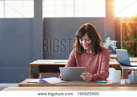 Attractive young businesswoman smiling while sitting alone at her desk in a modern office working online with a digital tablet