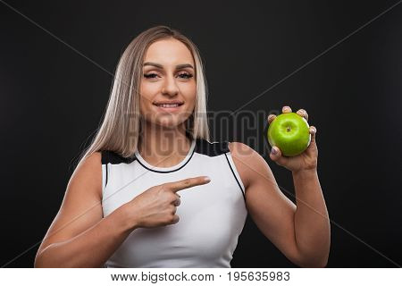 Sporty beefy blonde woman smiling wearing top pointing at green apple she holds.