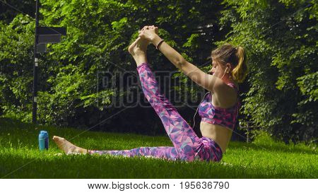 Young woman doing yoga exercises in the park. Akarna Dhanurasana