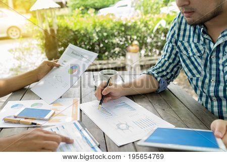 Close up young smart asian business man working on report chart debate consulting with co worker casual style at outdoor cafe on wooden table with coffee in the morning technology smartphone tablet concept. selective focus on hand and pen.