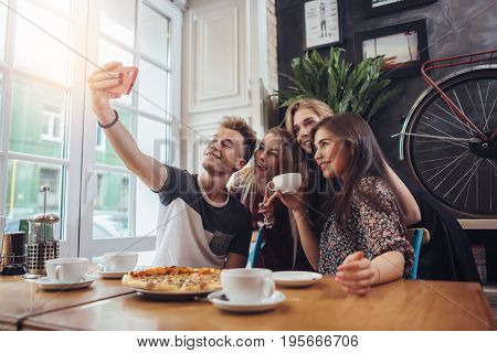 A guy taking a self-portrait with his female groupmates using his smartphone while hanging out in a coffee shop.