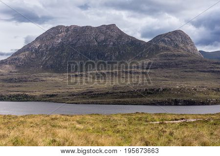 Assynt Peninsula Scotland - June 7 2012: Brown Mountain range along Loch Lurgainn under silver sky with gray clouds. Green vegetation on foothills and dry foreground.