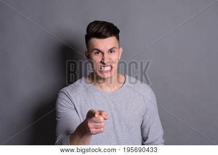 Feeling angry. Furious man grinting his theets and pointing with his finger to camera on grey studio background